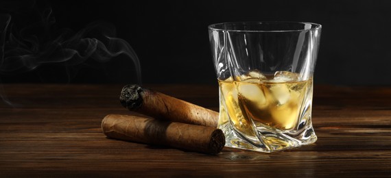 Glass of whiskey with ice cubes and cigars on wooden table