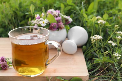 Cup of aromatic herbal tea, pestle and ceramic mortar with different wildflowers on green grass outdoors. Space for text