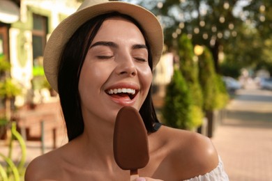 Photo of Beautiful young woman eating ice cream glazed in chocolate on city street, closeup