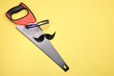 Photo of Man's face made of artificial mustache, safety glasses and hand saw on yellow background, top view. Space for text