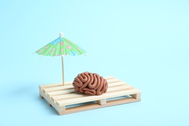 Photo of Brain made of plasticine on mini wooden sunbed under umbrella against light blue background, space for text