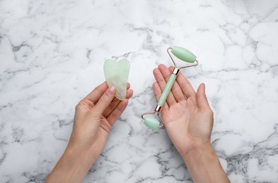 Photo of Woman holding gua sha stone and face roller at white marble table, top view