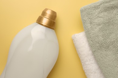 Photo of Bottle of fabric softener and towels on pale yellow background, flat lay