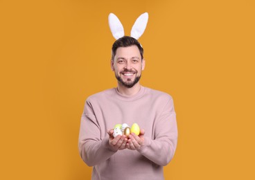 Photo of Happy man in bunny ears headband holding painted Easter eggs on orange background