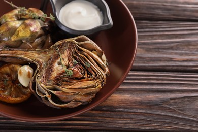 Tasty grilled artichoke and sauce in bowl on wooden table, closeup. Space for text