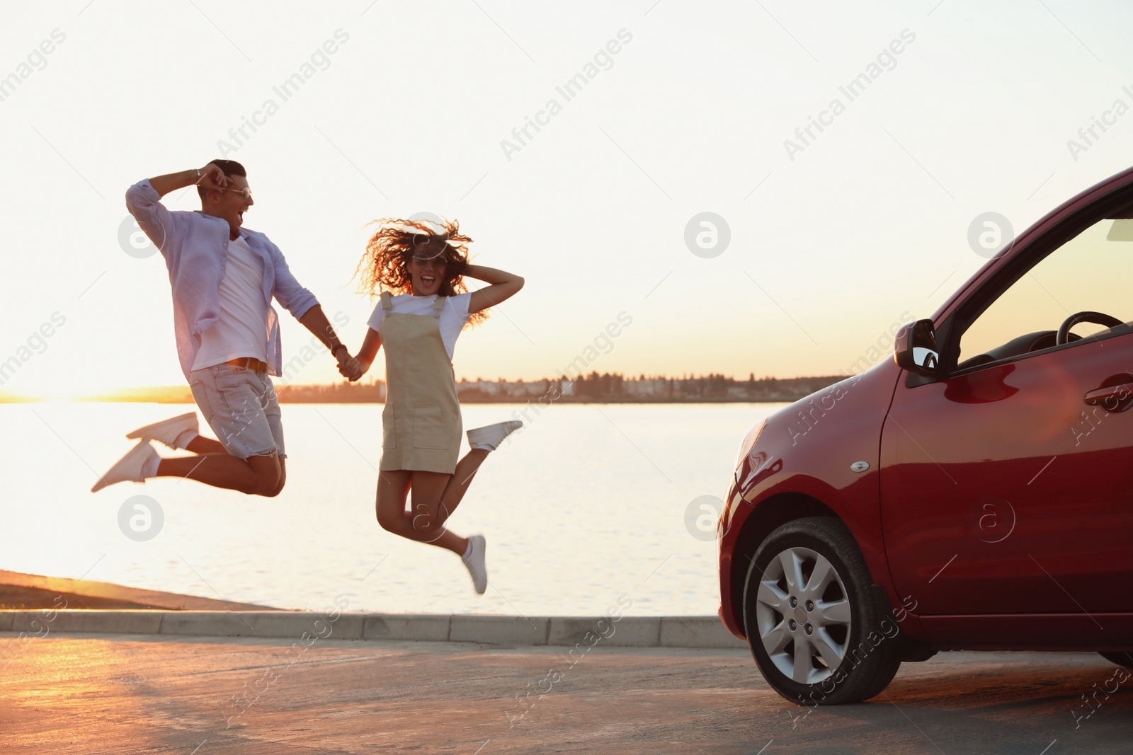 Photo of Happy couple jumping near car outdoors at sunset. Summer trip