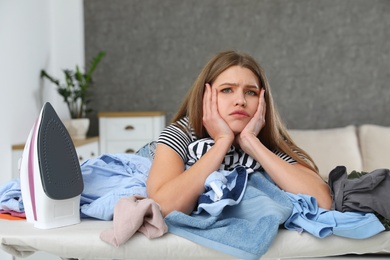 Photo of Tired woman leaning on ironing board with clothes at home