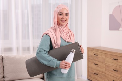 Photo of Muslim woman in hijab with fitness mat and bottle of water at home