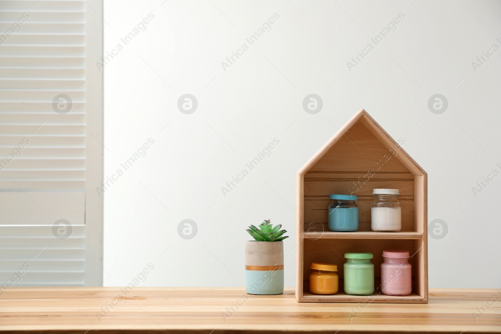 Photo of House shaped shelf with jars of paints and flower on table indoors. Space for text