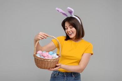 Photo of Easter celebration. Happy woman with bunny ears and wicker basket full of painted eggs on grey background