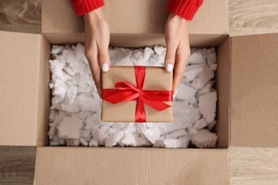 Woman putting Christmas gift box into parcel at wooden table, top view. Sending present by mail