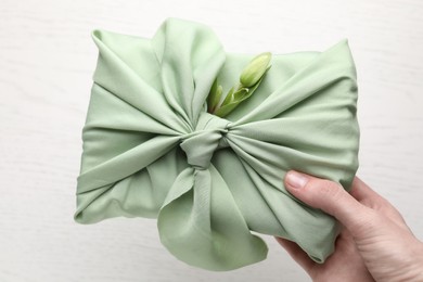 Photo of Furoshiki technique. Woman holding gift packed in green fabric with hellebore flower over light table, top view