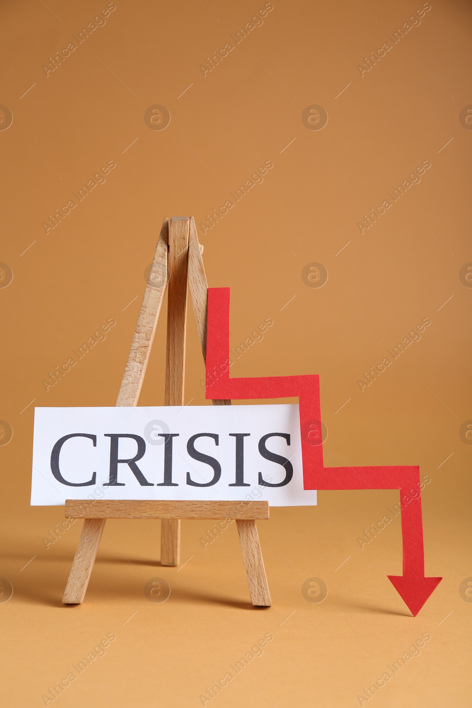 Photo of Wooden easel with word Crisis and descending red arrow on brown background
