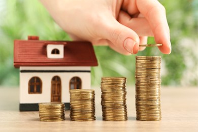 Mortgage concept. Woman stacking coins against blurred green background, closeup. House model on wooden table