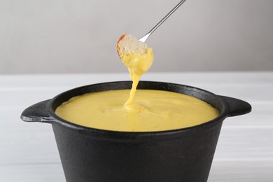 Dipping piece of bread into fondue pot with tasty melted cheese at white table, closeup