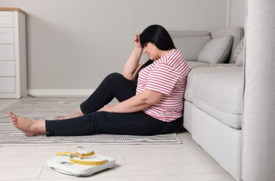 Photo of Depressed overweight woman sitting near sofa at home, focus on scales with measuring tape