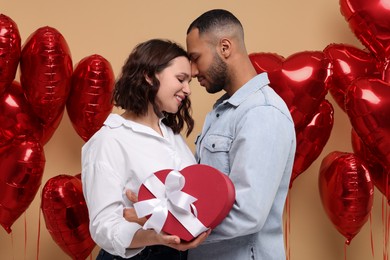 Photo of Lovely couple with gift box near heart shaped air balloons on beige background. Valentine's day celebration