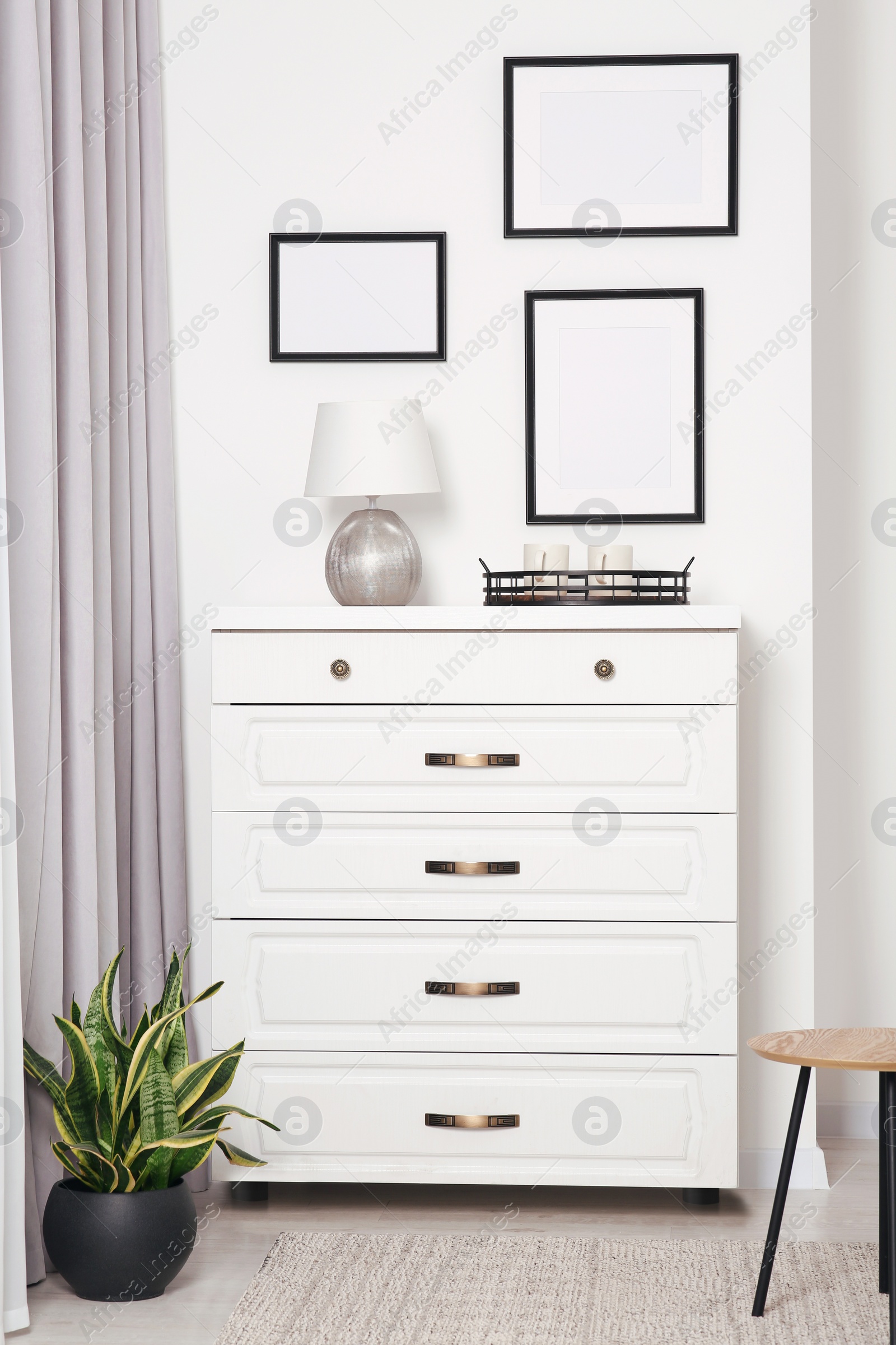 Photo of Empty frames hanging on white wall, chest of drawers and potted plant indoors