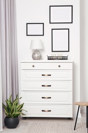 Photo of Empty frames hanging on white wall, chest of drawers and potted plant indoors