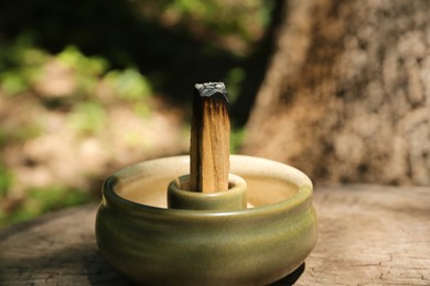 Photo of Smoldering palo santo stick in holder on wooden stump outdoors, closeup