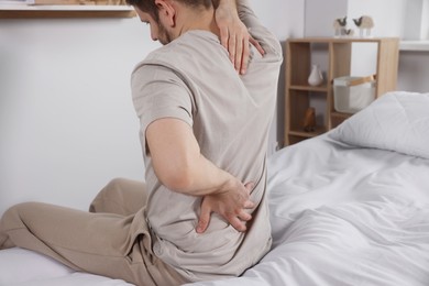 Photo of Man suffering from back pain while sitting on bed in room. Symptom of scoliosis