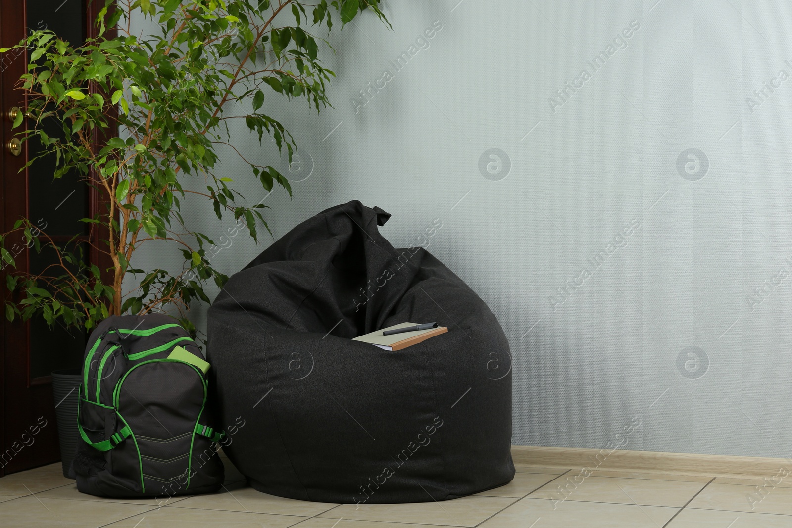 Photo of Black bean bag chair, houseplant and backpack near light grey wall in room. Space for text