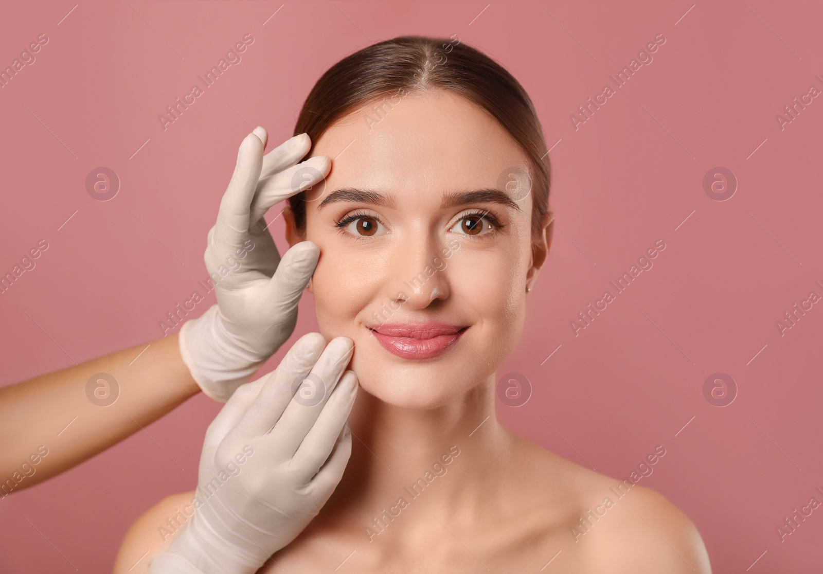 Photo of Doctor examining woman's face before plastic surgery on pink background