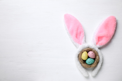 Headband with bunny ears, painted eggs and space for text on white wooden background, flat lay. Easter holiday