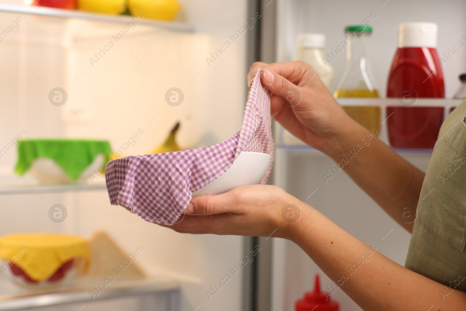 Photo of Woman taking away beeswax food wrap from bowl near refrigerator in kitchen, closeup