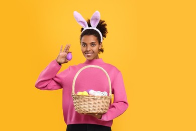 Happy African American woman in bunny ears headband holding wicker basket with Easter eggs on orange background. Space for text