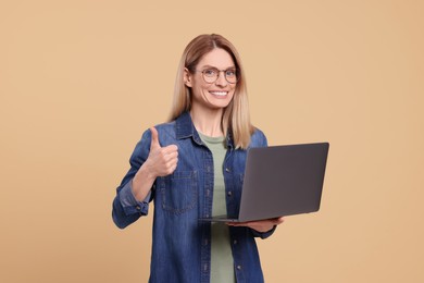 Happy woman with laptop showing thumb up on beige background