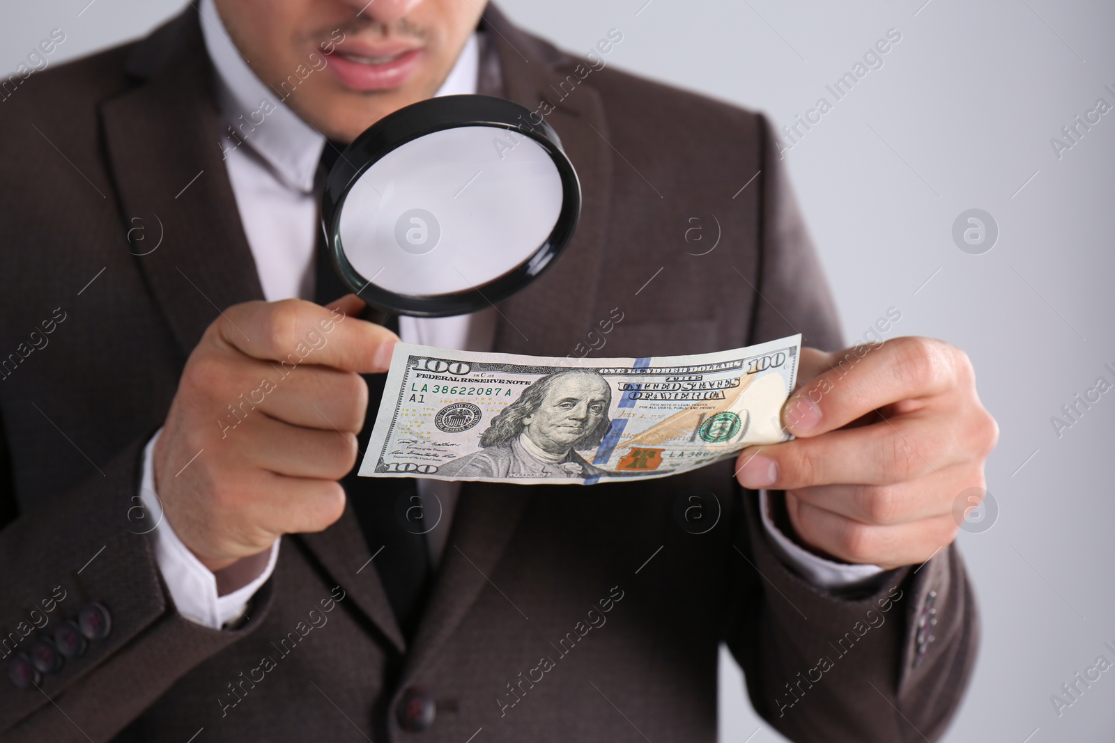 Photo of Expert authenticating 100 dollar banknote with magnifying glass on light grey background, closeup. Fake money concept