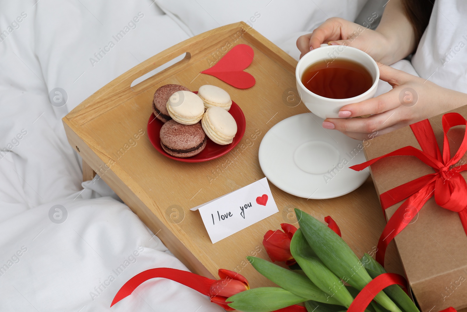 Photo of Tasty breakfast served in bed. Woman with tea, macarons, gift box, flowers and I Love You card at home, closeup