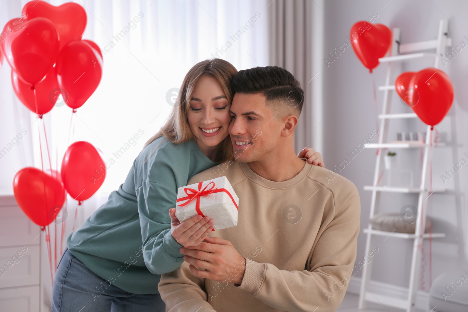 Photo of Woman presenting gift to her boyfriend in room decorated with heart shaped balloons. Valentine's day celebration