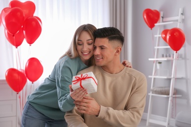 Woman presenting gift to her boyfriend in room decorated with heart shaped balloons. Valentine's day celebration