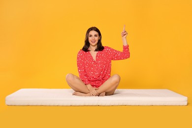 Young woman sitting on soft mattress and pointing upwards against orange background