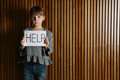 Sad little girl with sign HELP on wooden background, space for text. Child in danger