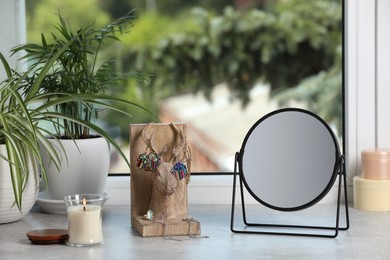 Mirror, jewelry, candles and houseplants on light grey stone window sill