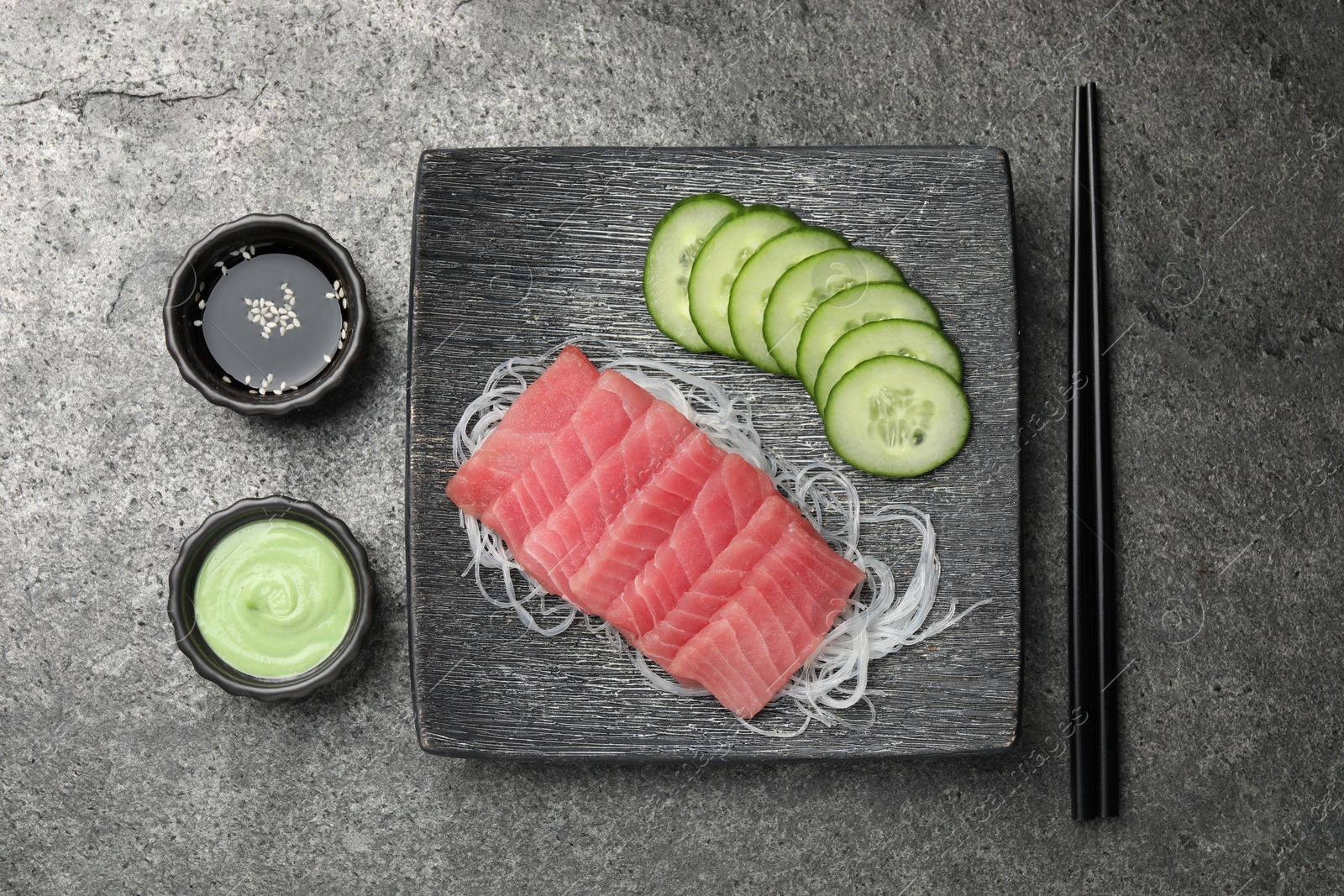 Photo of Tasty sashimi (pieces of fresh raw tuna), cucumber slices, glass noodles, soy sauce and wasabi on gray table, flat lay