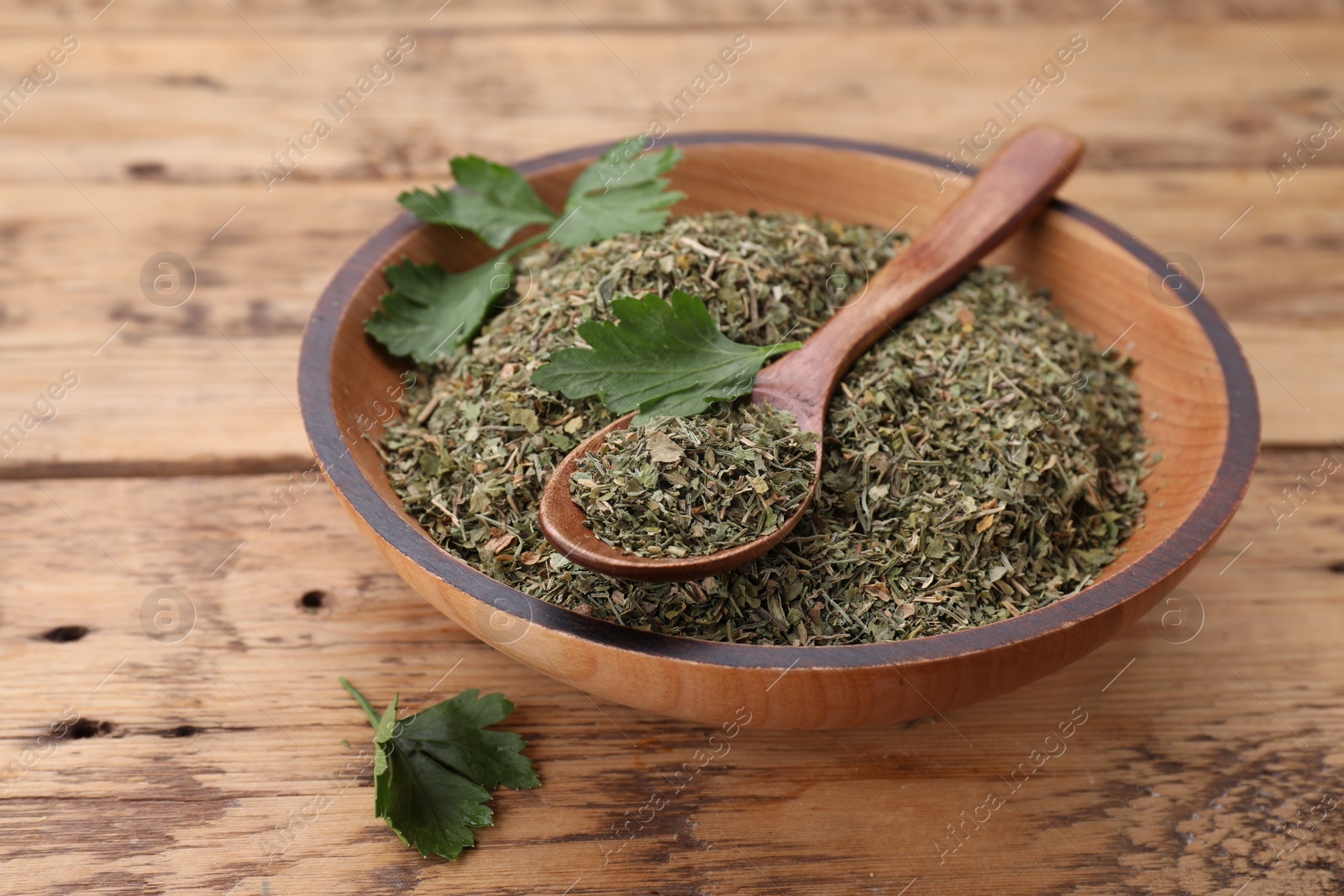 Photo of Bowl and spoon with dried parsley on wooden table, closeup