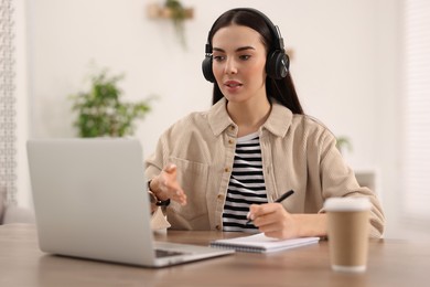 Photo of Young woman in headphones using video chat during webinar at table in room