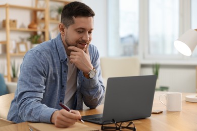 Photo of Happy man writing notes while working on laptop at wooden desk in room