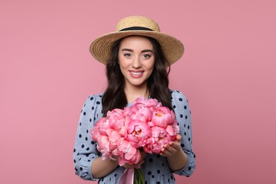 Photo of Beautiful young woman in straw hat with bouquet of peonies on pink background