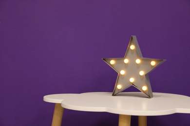 Photo of Stylish star shaped glowing night lamp on white table against purple background. Space for text