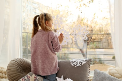 Photo of Little girl decorating window with paper snowflake indoors