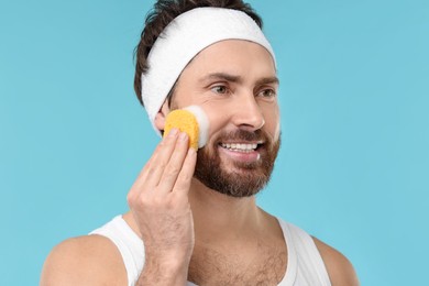 Man with headband washing his face using sponge on light blue background