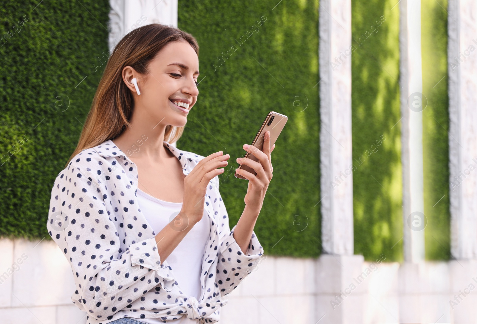 Photo of Young woman with wireless headphones and mobile device listening to music near green grass wall. Space for text