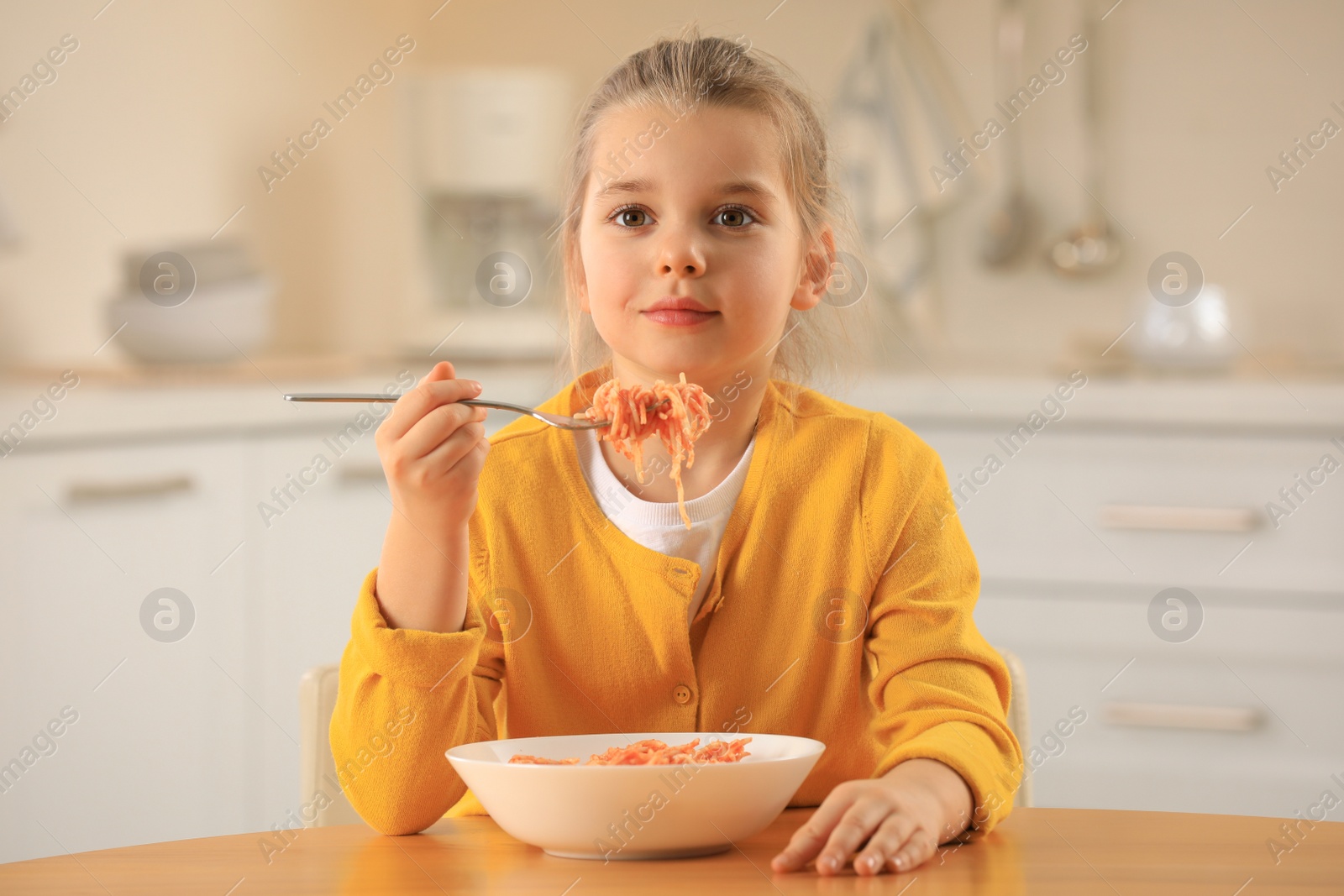 Photo of Cute little girl eating tasty pasta at table in kitchen