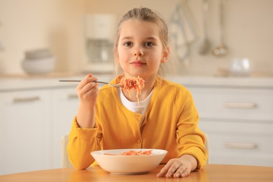 Photo of Cute little girl eating tasty pasta at table in kitchen