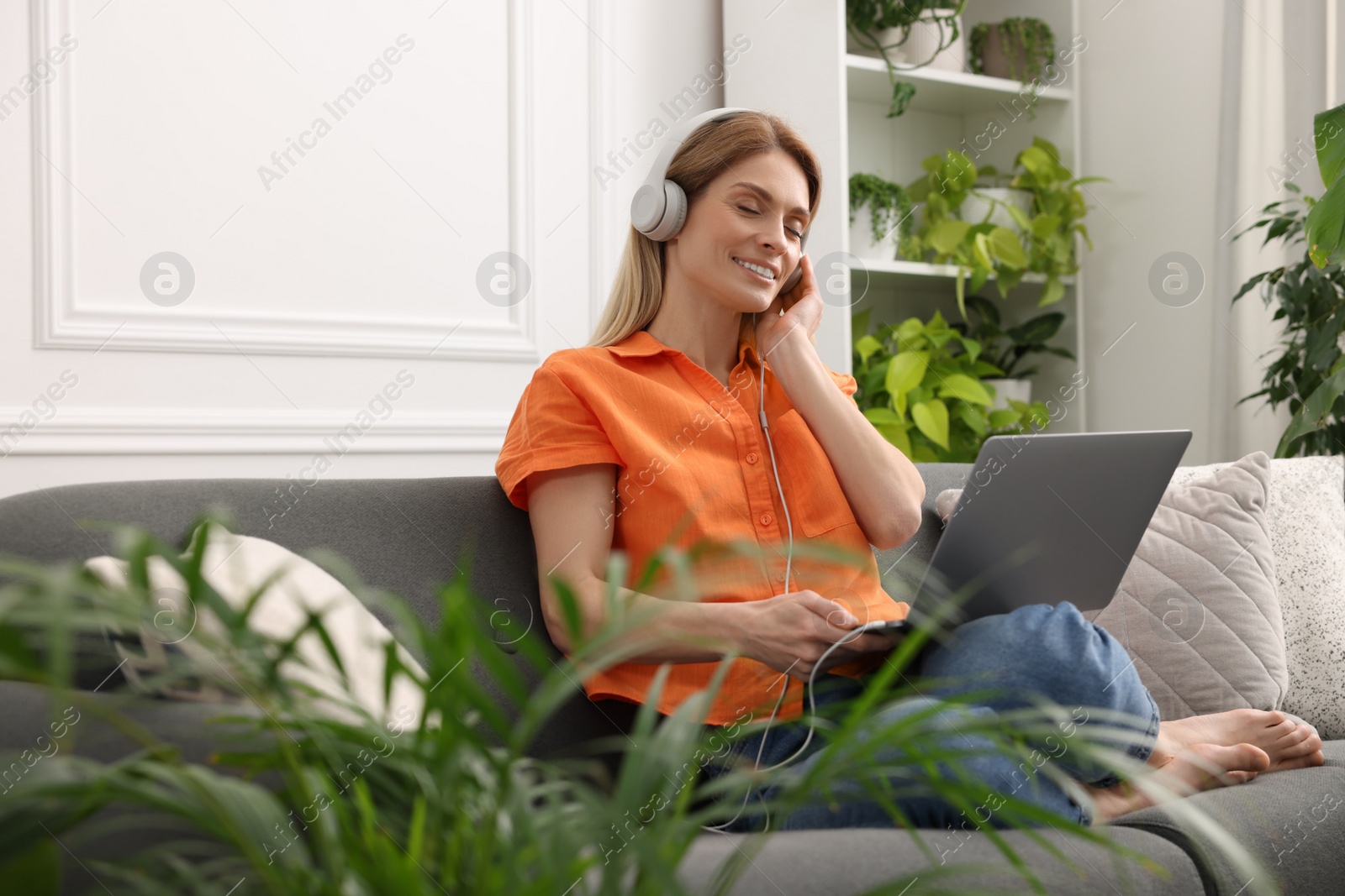 Photo of Woman in headphones using laptop on sofa near beautiful potted houseplants at home